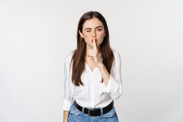 Smiling beautiful woman shushing, making hush shh sign, press finger to lips and winking, hiding secret, standing in blouse and jeans over white background