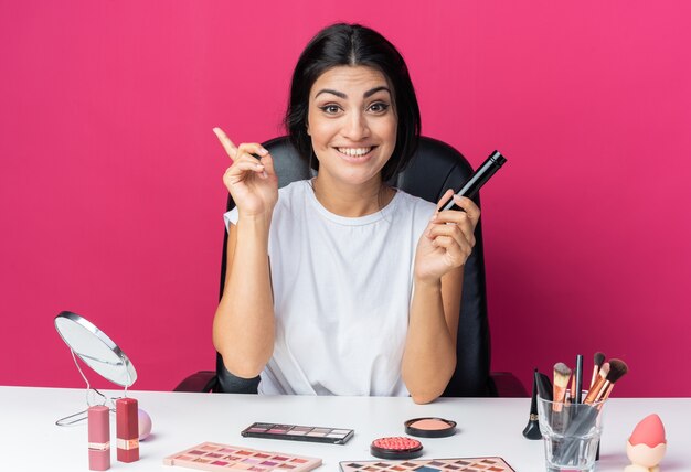 Smiling beautiful woman sits at table with makeup tools holding powder brush points at up 