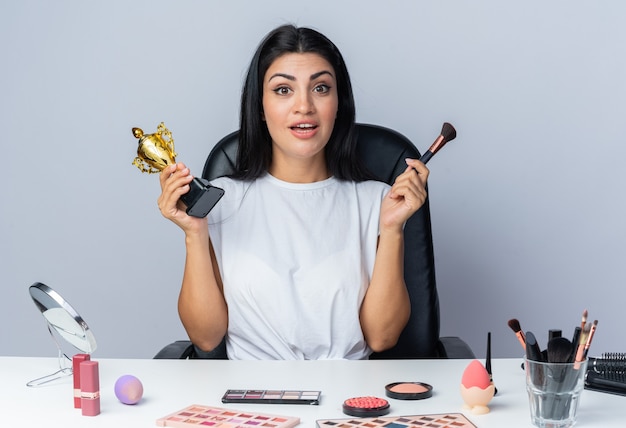 Smiling beautiful woman sits at table with makeup tools holding winner cup with powder brush 