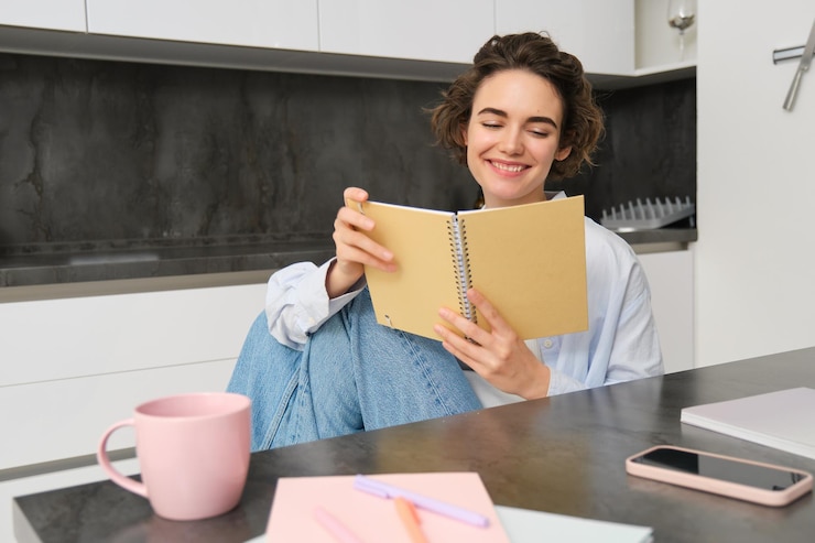 Free Photo smiling beautiful woman sitting with notebook in kitchen reading notes studying doing homework