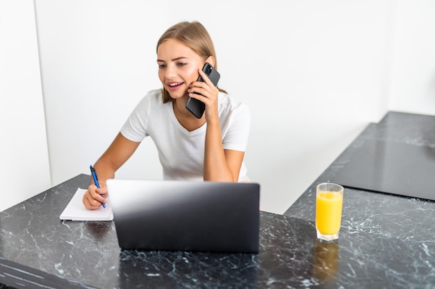 Smiling beautiful woman talking on mobile phone while sitting at the kitchen table with laptop and notepad