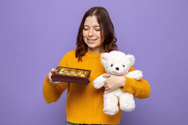 Улыбающаяся красивая молодая девушка в счастливый женский день держит плюшевого мишку, глядя на коробку конфет в руке, изолированной на синей стене