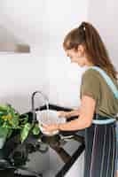 Free photo smiling beautiful young woman holding colander under the flowing water in the sink