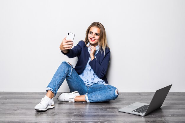 白い壁の床に座っている笑顔の美少女学生と彼女がラップトップコンピューターの研究を使用して幸せな携帯電話でビデオ通話。