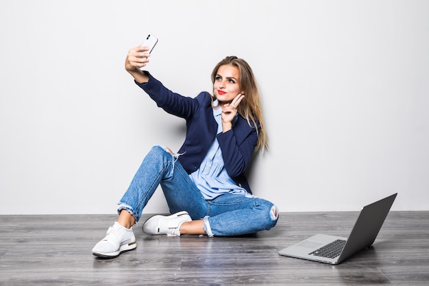 Улыбающийся студент-красотка сидит на полу с белой стеной и видеозвонком на мобильный телефон с счастливым, когда она использует исследование портативного компьютера.