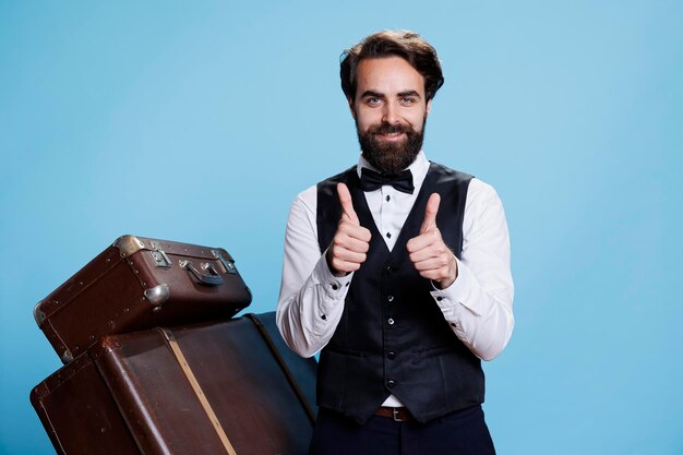 Smiling bellboy does thumbs up sign while he is wearing formal hotel attire in studio, expressing success and agreement. Young man dressed as doorman shows like symbol, tourism industry.