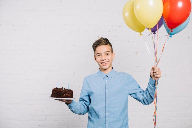 Smiling birthday boy holding balloons and chocolate cake standing against wall