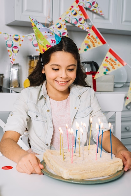 Free photo smiling birthday girl wearing party hat on head looking at cake decorated with colorful candles