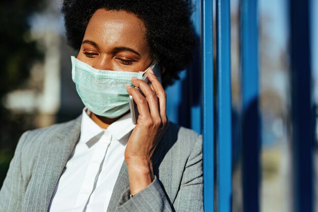 Улыбающаяся чернокожая деловая женщина общается по мобильному телефону в защитной маске на лице во время эпидемии вируса