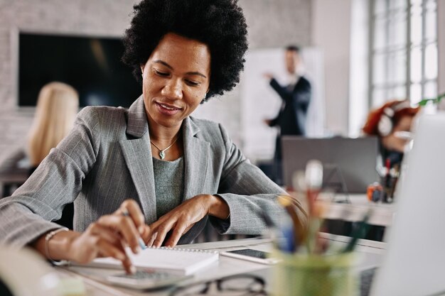 Smiling black entrepreneur working on financial reports at her desk in the office There are people in the background