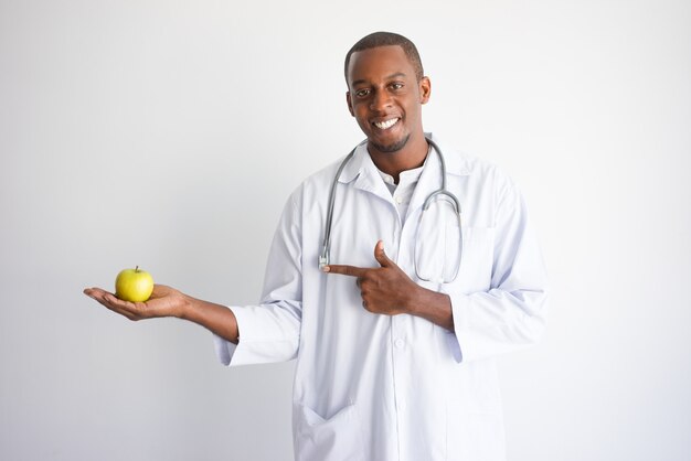 Улыбается черный мужчина-врач Холдинг и указывая на яблоко.