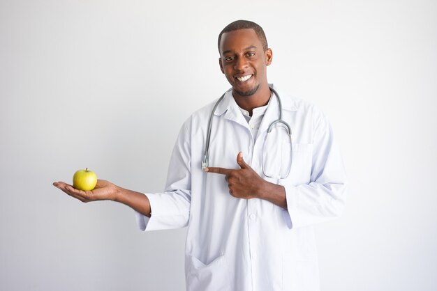 Smiling black male doctor holding and pointing at apple. 