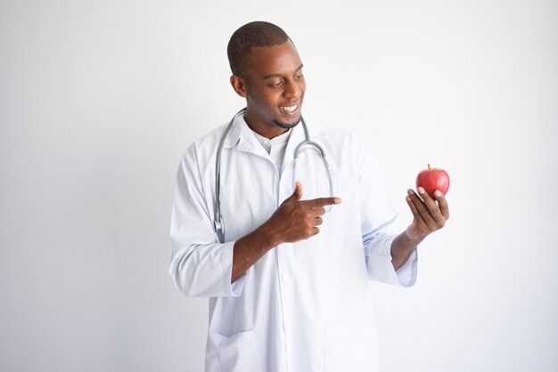 Улыбается черный мужчина-врач Холдинг и указывая на красное яблоко.