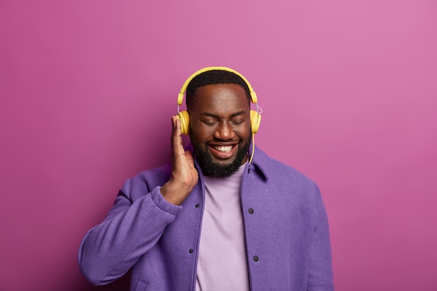 Smiling black man enjoys good sound in headphones, made new playlist, listens favourite music during spare time, dressed in purple jacket, shows white teeth. People