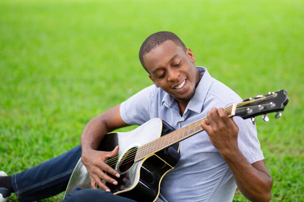 Улыбающийся черный человек, играющий на гитаре и сидящий на траве