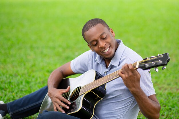 Smiling black man playing guitar and sitting on grass