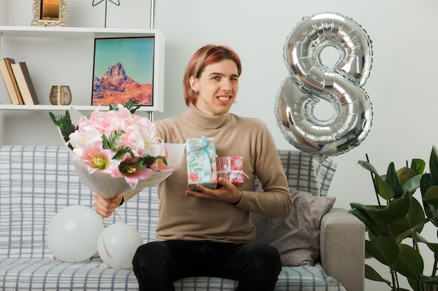Smiling blinked handsome guy on happy women day holding present with bouquet sitting on sofa in living room