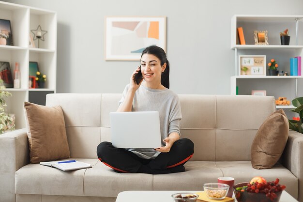 Smiling blinked young girl holding and used laptop speaks on phone sitting on sofa behind coffee table in living room