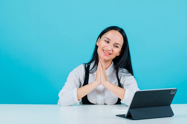 Smiling blogger girl is holding hands together by posing to tablet camera on blue background