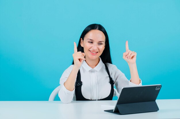 Smiling blogger girl is looking at tablet screen by pointing up with forefinger on blue background