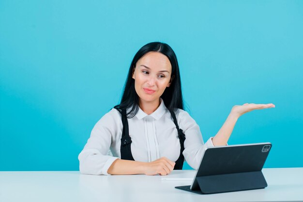 Smiling blogger girl is pointing right with hand by sitting in front of tablet camera on blue background