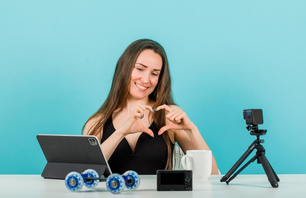 Smiling blogger girl is posing at little camera by showing heart gesture on blue background