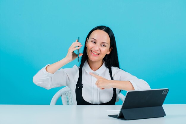 Smiling blogger girl is posing to tablet camera by holding phone on blue background