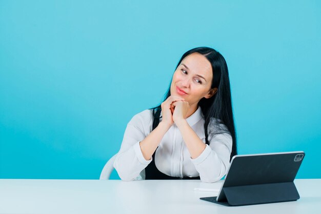 Smiling blogger girl is posing to tablet camera by putting hands on chin on blue background