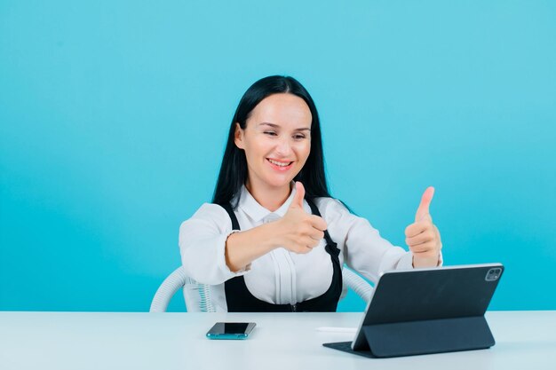 Smiling blogger girl is posing to tablet camera by showing perfect gesture on blue background