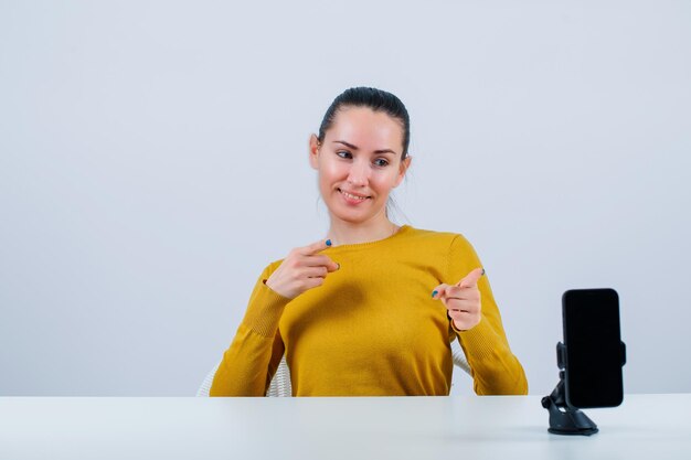 Smiling blogger girl is showing hand gesture by sitting in front of mobile camera on white background