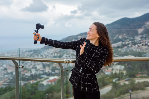 Smiling blogger girl is showing hi gesture to camera in her hand against the background of city view