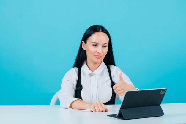 Smiling blogger girl is showing perfect gesture by looking at her tablet on blue background