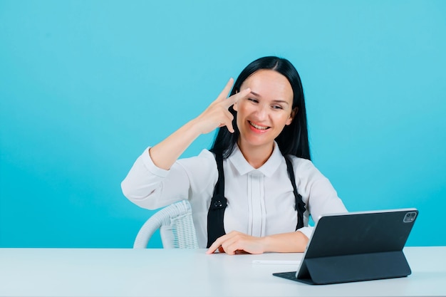 Smiling blogger girl is showing two gesture near eye by posing to tablet camera on blue background