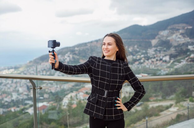 Улыбающаяся девушка-блогер делает селфи со своей камерой, положив руку на талию на фоне вида на город