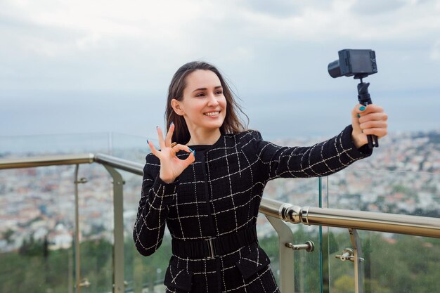 Smiling blogger girl is taking selfie with her mini camera by showing perfect gesture against the background of city view