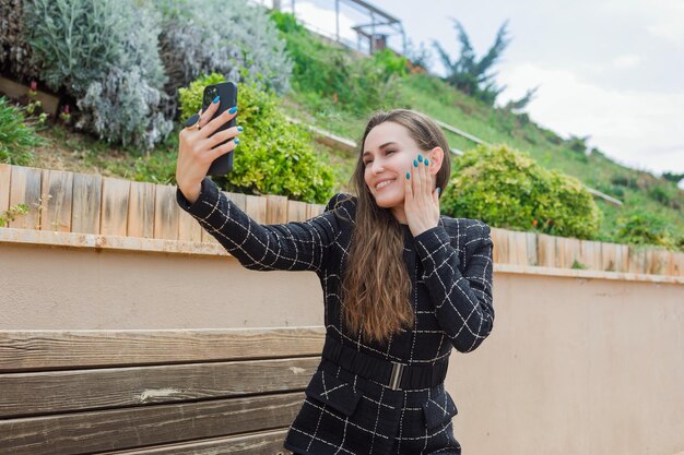 笑顔のブロガーの女の子が公園で頬に手をかざしてスマートフォンで自分撮りをしています