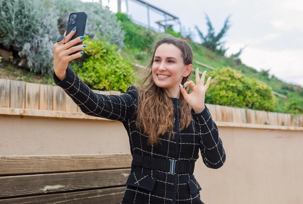 Smiling blogger girl is taking selfie with her smartphone by showing okay gesture in park