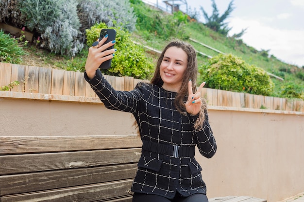 無料写真 笑顔のブロガーの女の子が公園で勝利のジェスチャーを示すことによって彼女のスマートフォンで自分撮りを取っています