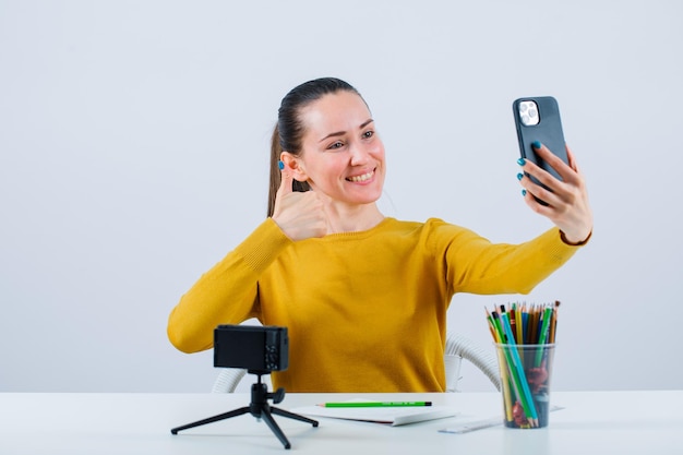 Smiling blogger girl is taking selfie with phone by showing perfect gesture on white background