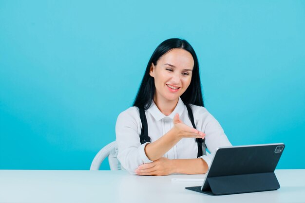 Smiling blogger girl is talking on video call by extending hand to tablet camera on blue background