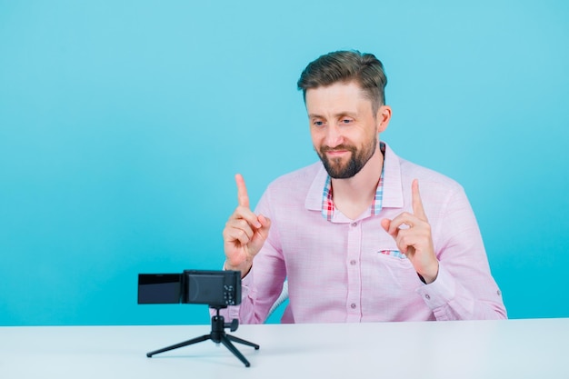 Smiling blogger man is posing to his mini camera by pointing up with forefingers on blue background