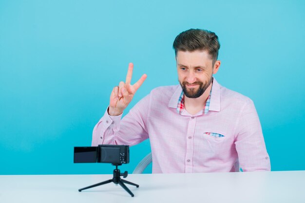 Smiling blogger man is posing to his mini camera by showing two gesture and sitting on blue background