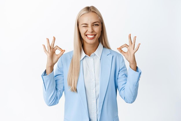 Smiling blond businesswoman showing okay signs winking and looking satisfied approve and praise smth good wearing corporate clothing white background