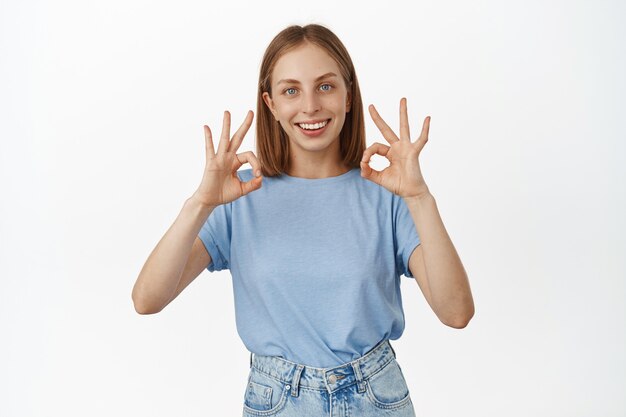Smiling blond caucasian woman shows okay OK signs, approve something cool, praise great job, recommending product, standing over white wall