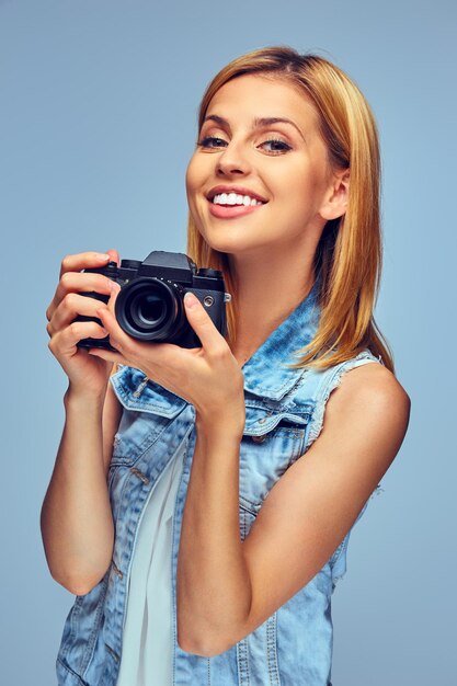 Улыбающаяся блондинка держит компактный цифровой фотоаппарат, изолированный на сером фоне.
