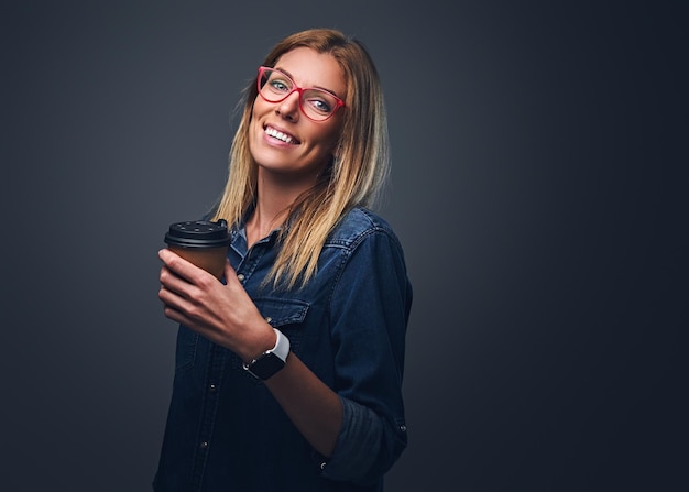 Smiling blond female in red eyeglasses holds take away coffee cup over grey background.