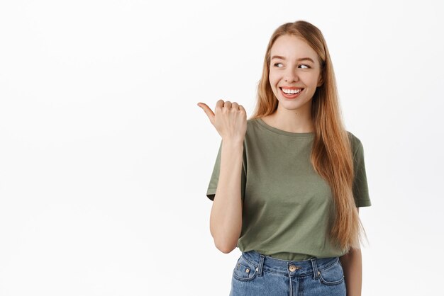 Smiling blond girl looks at left side and points at product reads promotional text aside on white copy space wears summer clothes against white background