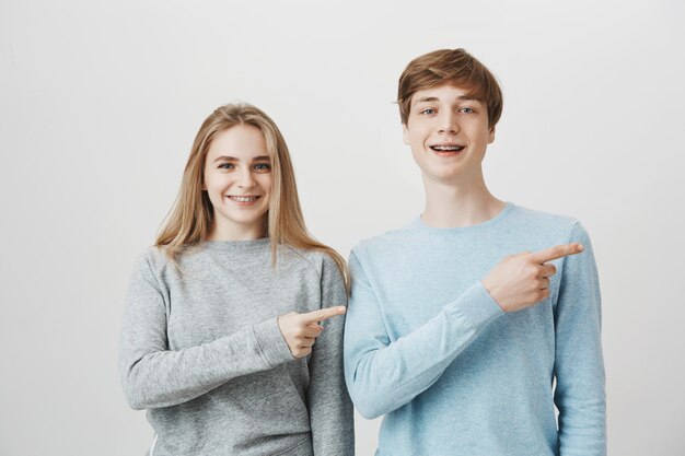 Smiling blond guy and girl with braces showing way, pointing fingers right