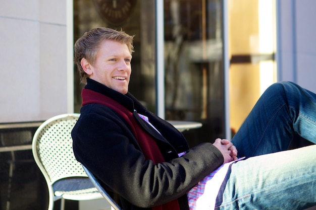 Smiling blond male in a black coat sitting on a chair in the street