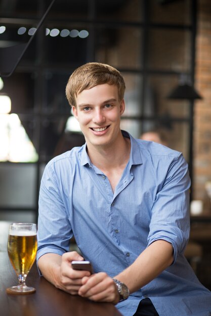 Улыбающийся блондин с пивом и смартфоном в баре